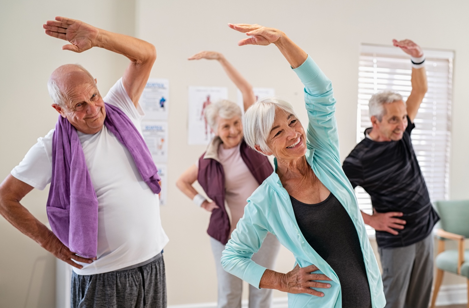 A group of seniors in an exercise class smiling and stretching with one hand over their heads.