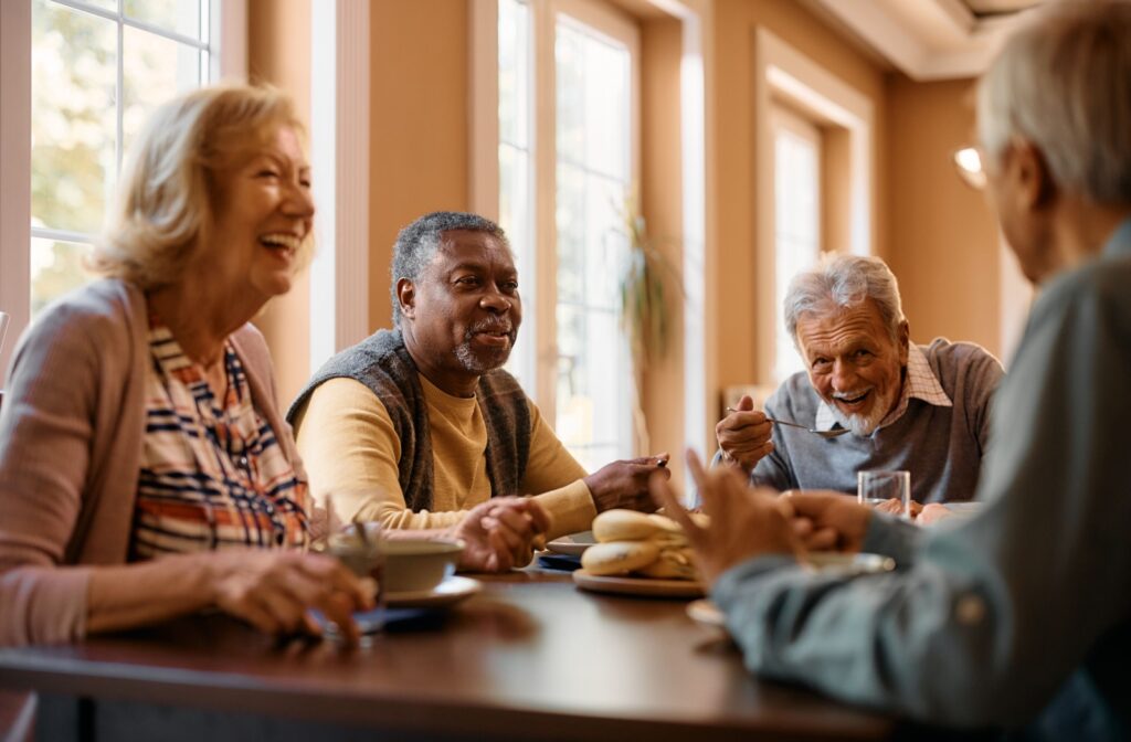 Happy seniors talk and laugh while eating at a dining table in a senior living community.