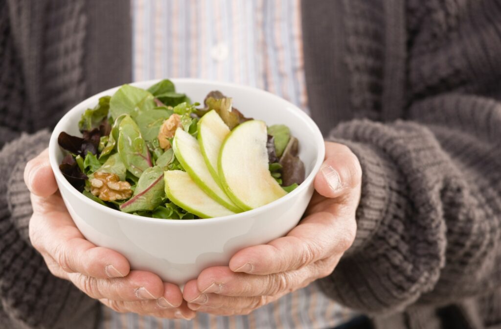 A senior wearing a grey cardigan holds a bowl full of leafy greens, nuts, and fruit for lunch to promote cognitive health