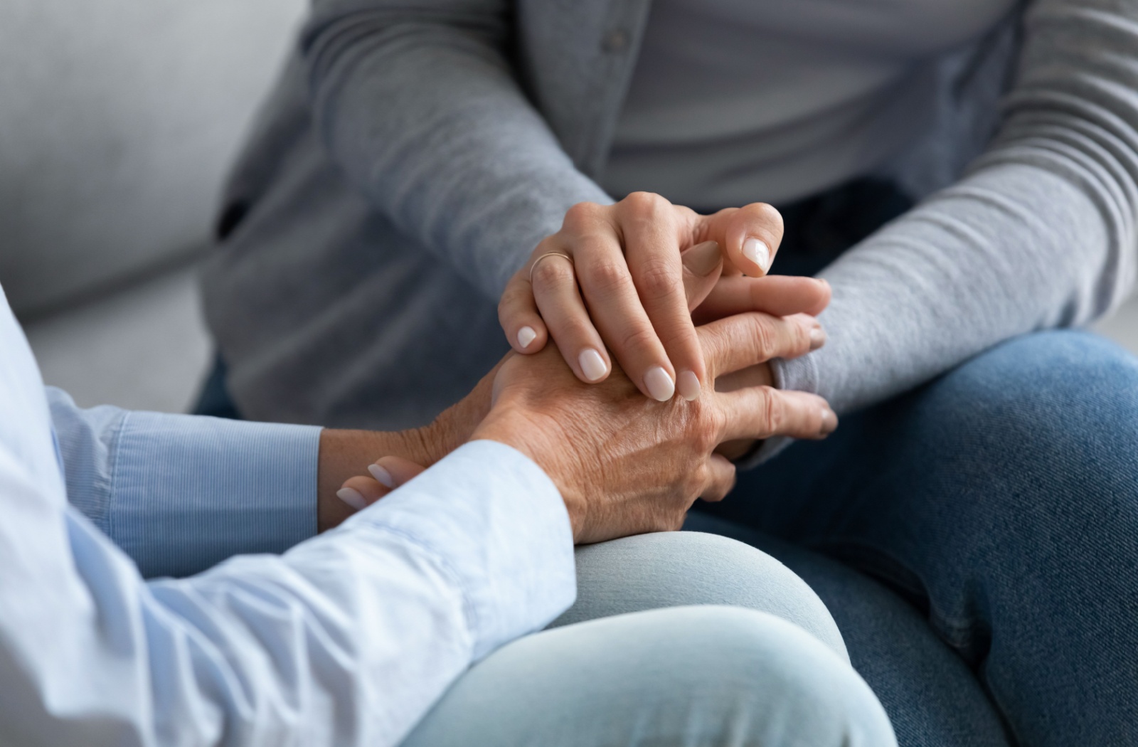 An adult child clasps their older parent's hands while sitting on a couch together during a serious conversation about assisted living