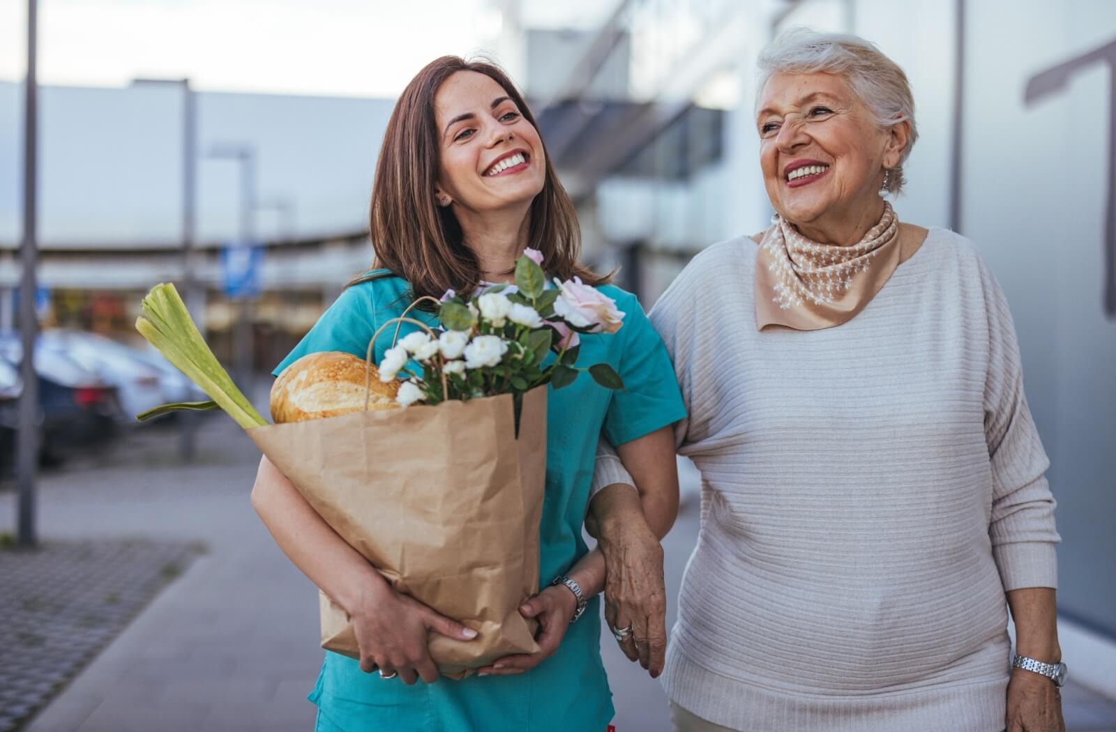 An older adult and a caregiver carry a bag of groceries and walk beside a senior living community while smiling