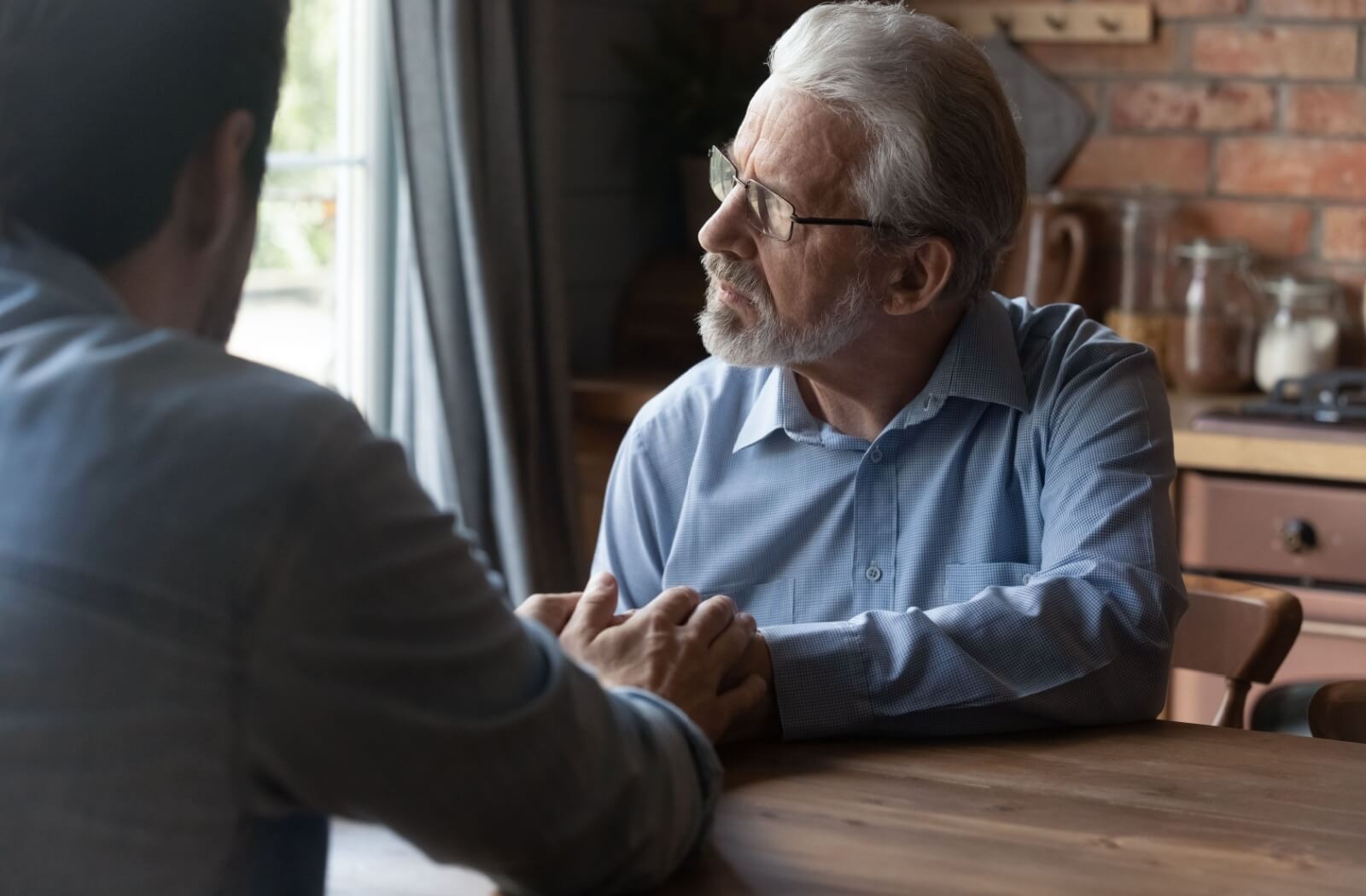 An older adult thoughtfully looks out the window while their adult child holds their hands during a talk in the dining room about moving to assisted living
