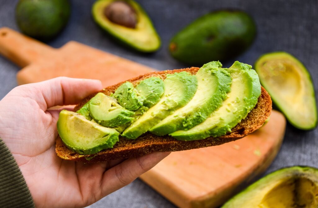 Avocado slices spread on toasted bread with more ripe avocados in the background.