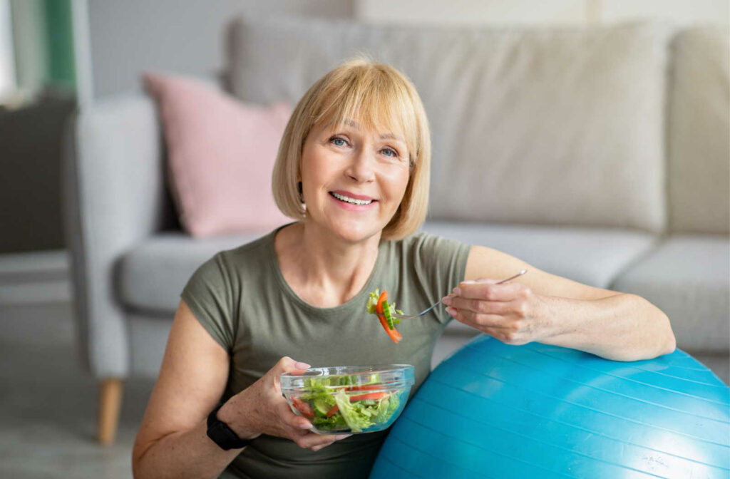 Senior woman eating a healthy salad while sitting next to an exercise ball.