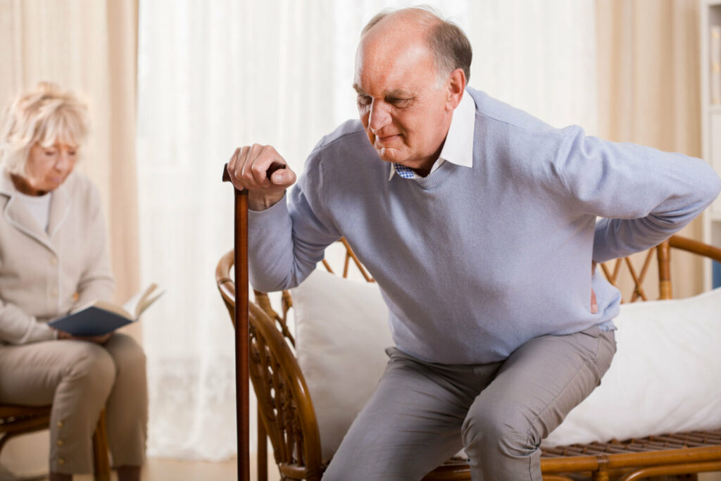 An elderly man is having a hard time standing from a sitting position. Holding his hips while standing