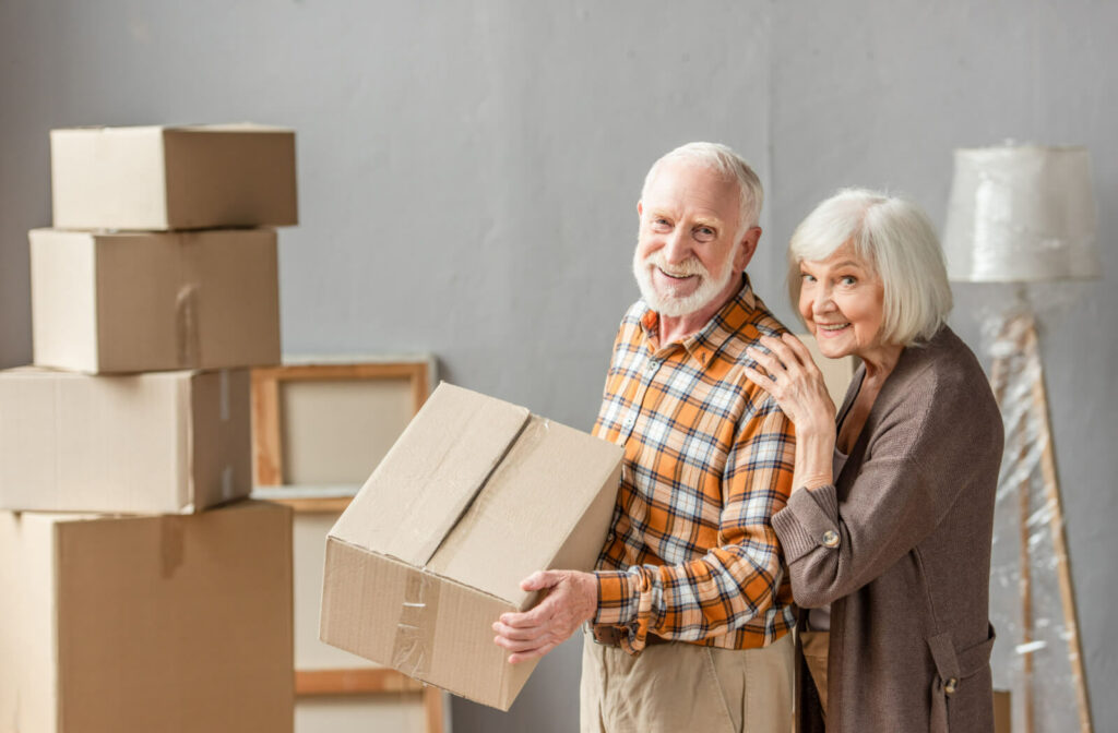 A senior man holding a cardboard box while his wife stands beside him. They are both smiling.