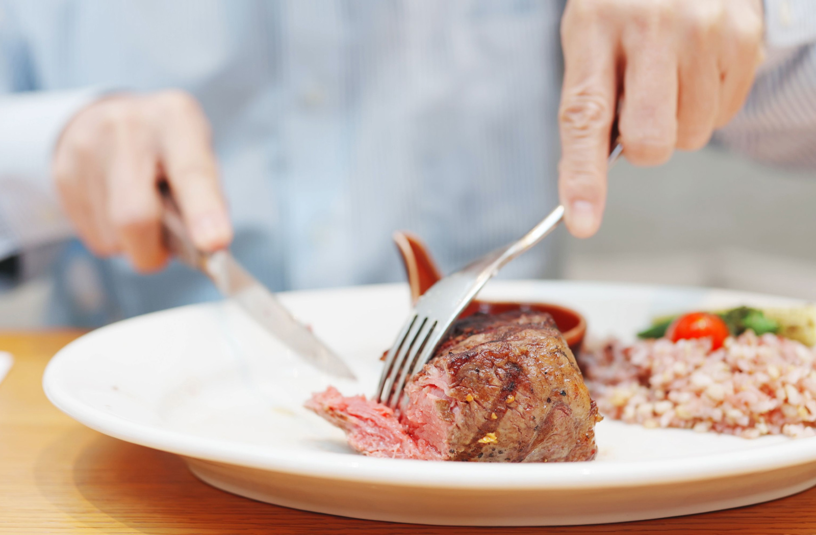 Close up of a senior man’s hand cutting a grilled steak with rice on the side of the plate.