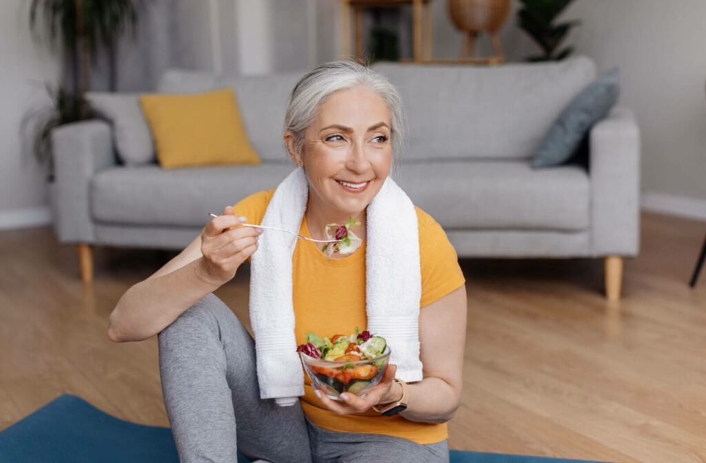 An active older adult sits on a yoga mat and enjoys a healthy salad after a workout.