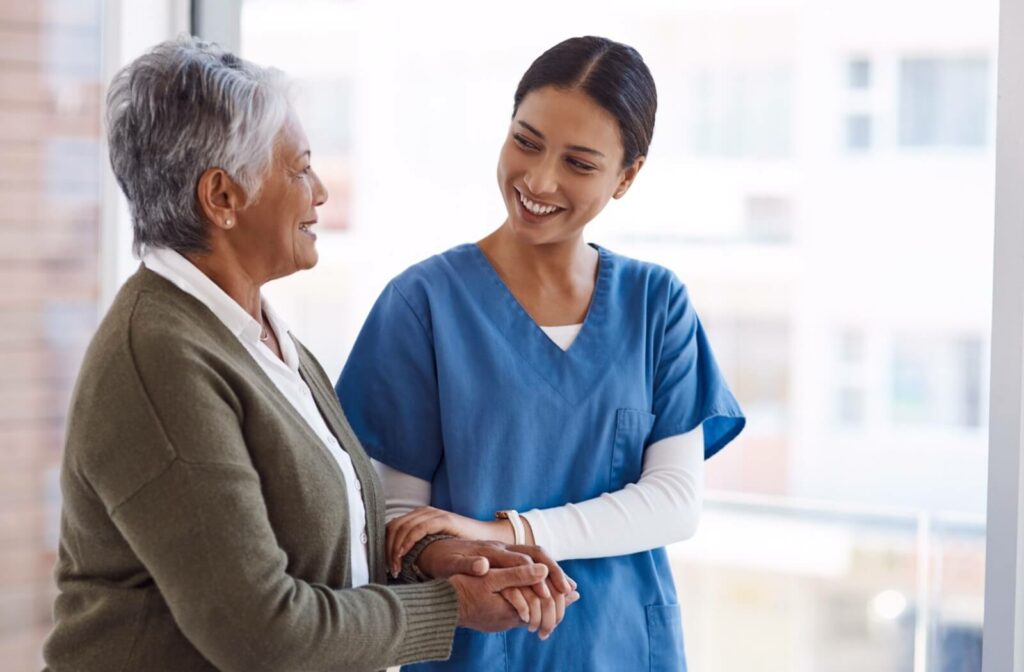 A senior woman and a caregiver arm-in-arm while smiling at each other walking together in assisted living.