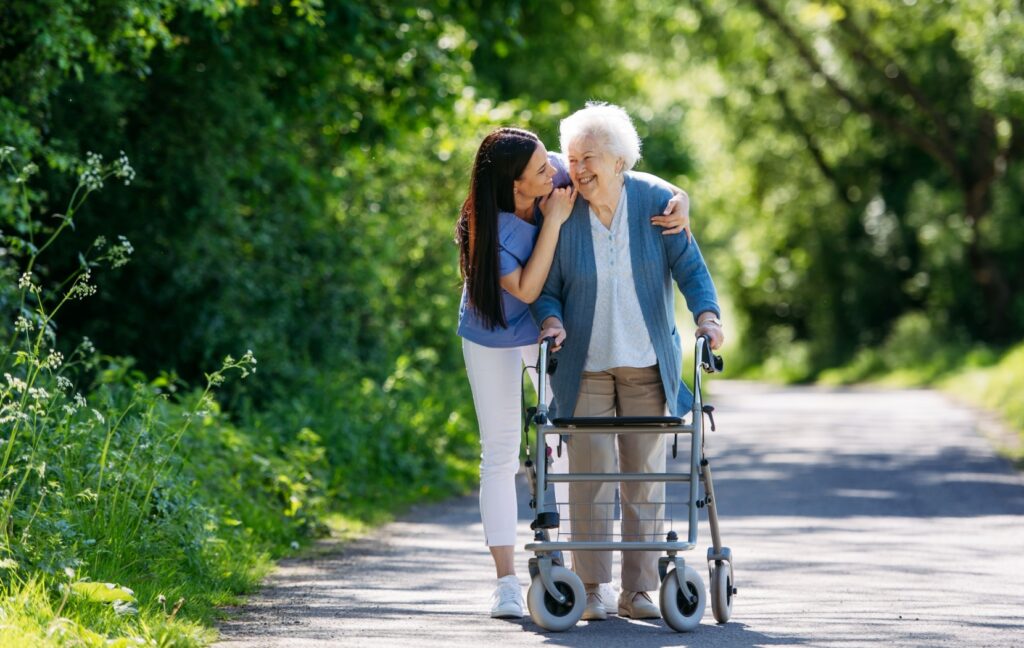 A senior with a walker and their caregiver walk down a paved pathway outside of a senior living community.