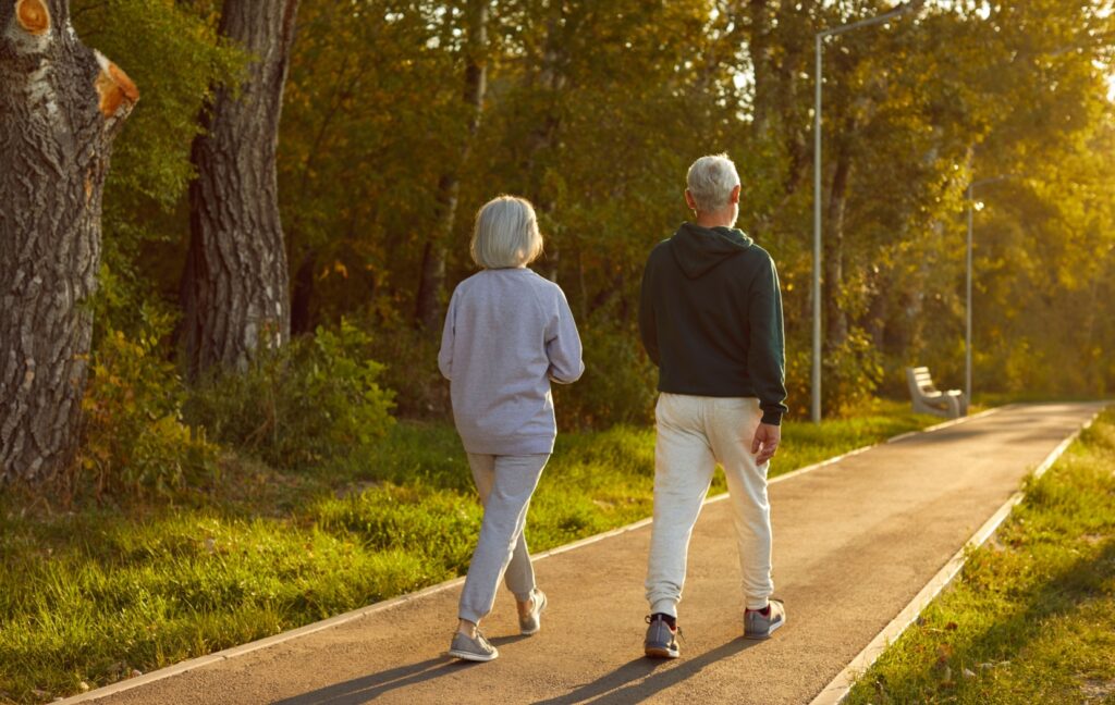 A senior couple walks on a path in a green park at sunset as the golden sun lights up the path ahead of them.