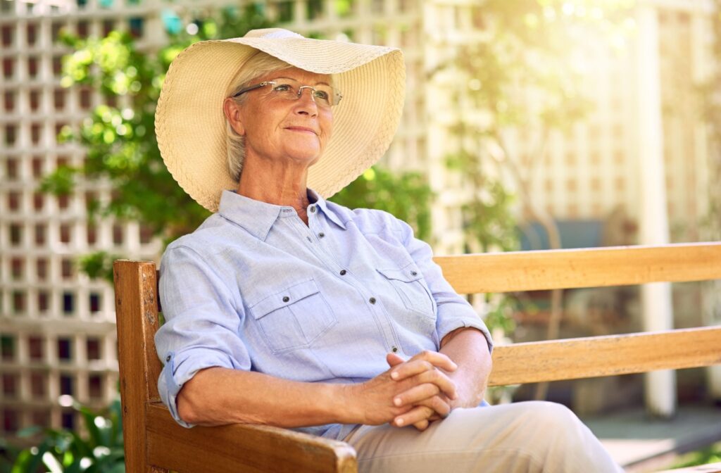 Person wearing a sunhat sits peacefully on a bench outdoors enjoying the sunlight in a quiet garden space.