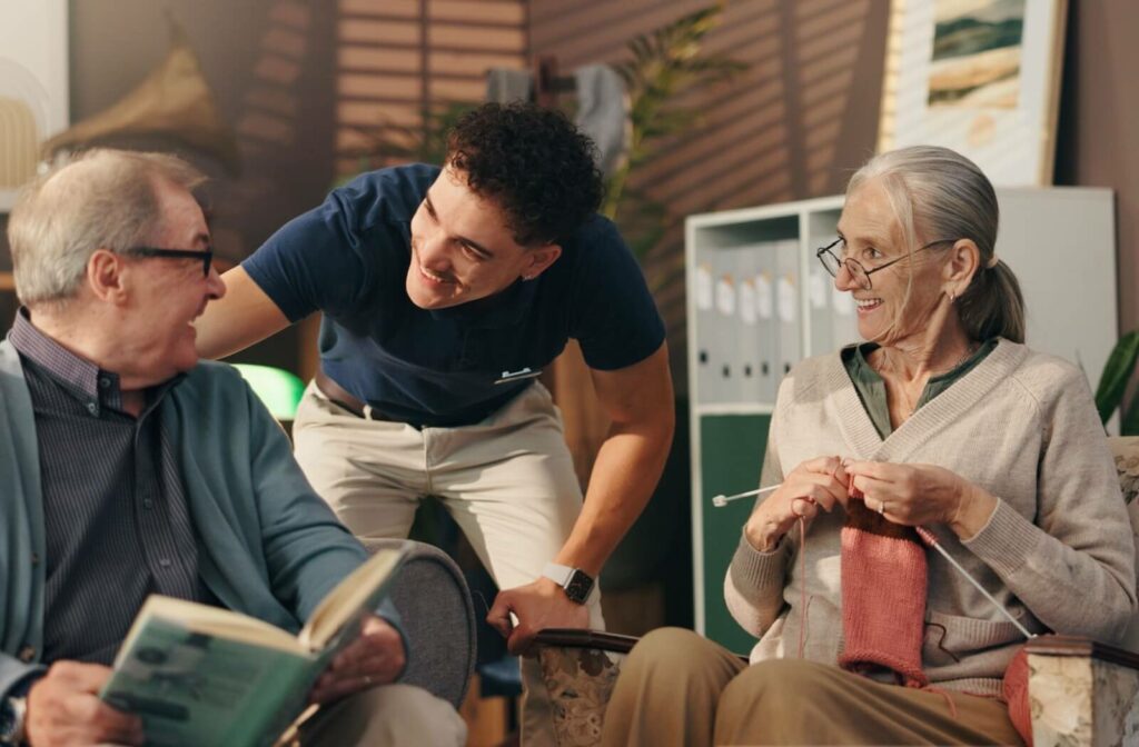 A smiling care team member interacts with two older adults, one knitting and the other reading, in a cozy community space.