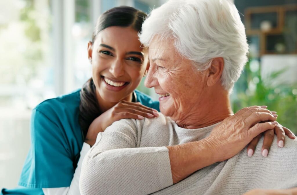 Smiling caregiver in blue scrubs with arm around senior woman in bright common area